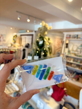 Hand holding a clear package with colorful fused glass fish holiday ornament in a store setting.
