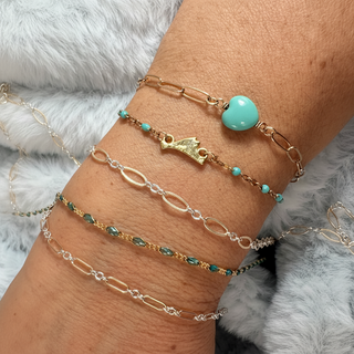 Multiple permanent bracelets on a wrist including a turquoise glass heart, martha's vineyard island shape and 2-tone chain, with a gray faux fur background