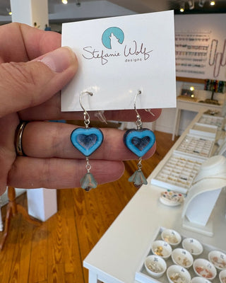 Heart-shaped earrings held in front of a branded card with a blurred store background